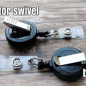 Puede incluir: Dos carretes retr&aacute;ctiles de credenciales negros con clips giratorios de cocodrilo y clips de cintur&oacute;n. Los carretes est&aacute;n sobre una superficie de madera.