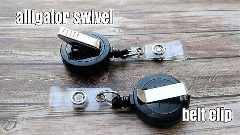 Puede incluir: Dos carretes retr&aacute;ctiles de credenciales negros con clips giratorios de cocodrilo y clips de cintur&oacute;n. Los carretes est&aacute;n sobre una superficie de madera.