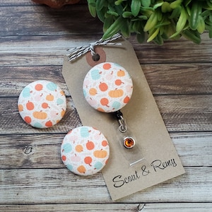 May include: Three fabric buttons with a pumpkin pattern on a white background. The buttons are attached to a brown card with the text "Scout & Remy".