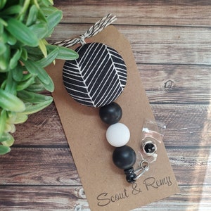 May include: Black and white patterned fabric circle with black and white beads attached to a clear badge holder with a metal clip. The badge holder has the text "Scout & Remy" printed on it.