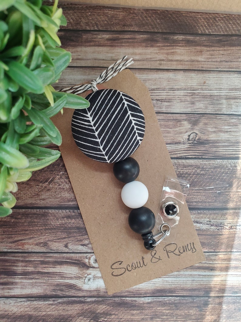 May include: Black and white patterned fabric circle with black and white beads attached to a clear badge holder with a metal clip. The badge holder has the text "Scout & Remy" printed on it.