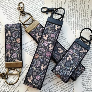 May include: Three fabric key fobs featuring a dark floral pattern with small winged creatures. Each strap is secured with a metal clamp and includes a swivel lobster clasp and a split ring. The hardware finishes include antique brass and matte black.
