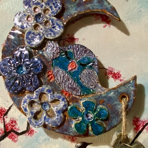 May include: A ceramic crescent moon ornament with blue flowers and an owl. The moon is glazed in shades of blue and brown, with a textured surface. The owl has blue and purple feathers. A black bird sits on a branch.