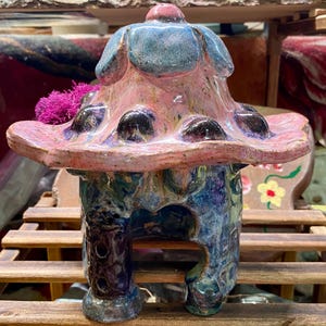 May include: A whimsical ceramic mushroom house sculpture. The house features a pink and blue cap with dark spots, a blue and purple base, and a dark brown chimney. A cluster of pink moss sits on the side. The sculpture is on a wooden surface.