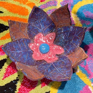 May include: A ceramic dish shaped like a flower with five petals. The petals are a deep purple with brown veins and a pink flower in the center with a blue center.