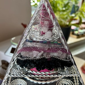 Könnte beinhalten: Eine klare Kristallpyramide mit Schichten aus verschiedenen Kristallen und Draht. Die Pyramide ist gefüllt mit rosa, weißen und schwarzen Kristallen und silbernem Draht.