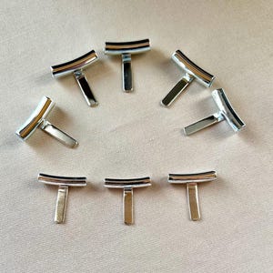 Könnte beinhalten: Eine Ansammlung von acht silberfarbenen Metallknebeln, die jeweils aus einem gebogenen horizontalen Stab bestehen, der an einem senkrechten, flachen rechteckigen Stift befestigt ist. Die Teile haben eine glatte, reflektierende Oberfläche.
