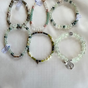 Peut inclure: Six bracelets en perles vertes et blanches avec des accents argentés. Les bracelets ont différents charmes, dont un arbre de vie, une perle argentée et un petit charme argenté. Les bracelets sont sur un fond blanc.