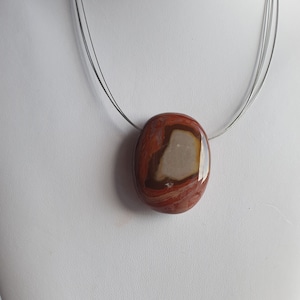 Op de afbeelding: Een gepolijste stenen hangerketting. De hanger heeft een ovale vorm met een mix van rode, bruine en crèmekleurige kleuren. De ketting heeft een meerstrengs zilverkleurige draad. De ketting wordt gepresenteerd op een witte mannequin.