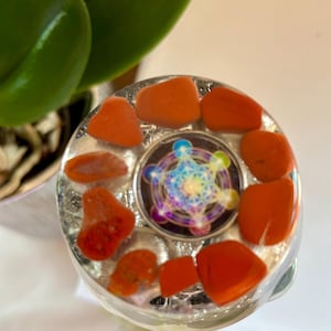 Könnte beinhalten: Eine durchsichtige Glasflasche mit einem silbernen Deckel und einem runden Aufkleber mit einem bunten geometrischen Muster. Die Flasche ist mit Wasser und orange-roten Steinen gefüllt.