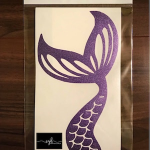 May include: A purple glitter mermaid tail decal inside a clear plastic sleeve. The decal features a cutout scale pattern on the tail and a fin design. A black rectangular logo with the text cgd and the URL https://m.facebook.com/CreativeGiftsandDecor is visible at the bottom.