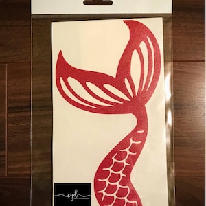 May include: A red glittery mermaid tail decal on a white backing sheet inside a clear plastic sleeve. The decal features a stylized fin and scale pattern. A black rectangular logo with the text cgd Creative Gifts and Decor is visible.