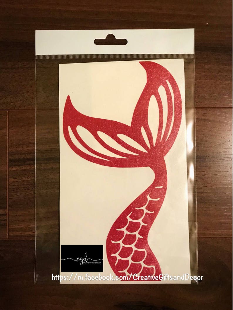 May include: A red glittery mermaid tail decal on a white backing sheet inside a clear plastic sleeve. The decal features a stylized fin and scale pattern. A black rectangular logo with the text cgd Creative Gifts and Decor is visible.