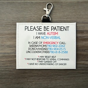 May include: A white tag with the text "PLEASE BE PATIENT I HAVE AUTISM I AM NON-VERBAL" and emergency contact information. The tag has a silver clip at the top.