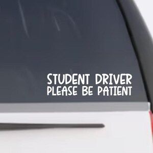 Peut inclure: Un autocollant en vinyle blanc appliqué sur une vitre de véhicule, présentant le texte "STUDENT DRIVER PLEASE BE PATIENT" dans une police sans empattement arrondie et grasse. Le lettrage est disposé sur deux lignes, centré sur la vitre.