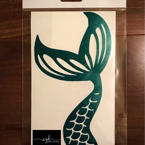 May include: A teal glitter mermaid tail decal on a white backing sheet inside a clear plastic sleeve. The decal features a scale pattern on the lower section and a cut-out fin design at the top. A small black logo with the text cgd Creative Gifts and Decor is visible at the bottom left.