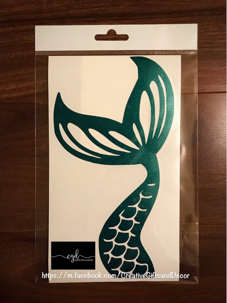 May include: A teal glitter mermaid tail decal on a white backing sheet inside a clear plastic sleeve. The decal features a scale pattern on the lower section and a cut-out fin design at the top. A small black logo with the text cgd Creative Gifts and Decor is visible at the bottom left.