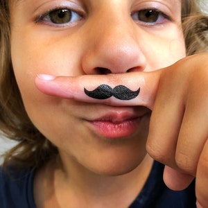 Puede incluir: Un tatuaje temporal de un bigote negro aplicado sobre la piel de un dedo. El diseño presenta una forma de bigote clásico con extremos curvados, realizado en tinta negra sólida y opaca que se sitúa horizontalmente sobre el dedo.