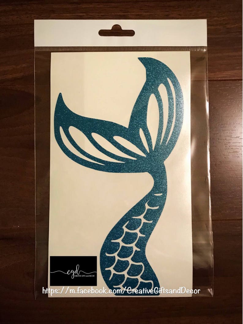 May include: A teal glitter mermaid tail decal on a white backing sheet inside a clear plastic sleeve. The design features a stylized fin and scale pattern. A small black rectangular logo with the text cgd Creative Gifts and Decor is visible.