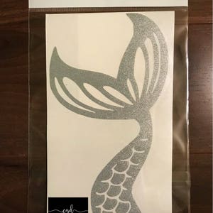 May include: A silver glitter mermaid tail decal enclosed in a clear plastic bag. The decal features a detailed scale pattern on the lower section and a wide, curved fin at the top. A black rectangular logo with the text cgd Creative Gifts and Decor is visible at the bottom left, along with the URL https://m.facebook.com/CreativeGiftsandDecor.