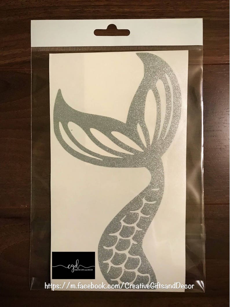 May include: A silver glitter mermaid tail decal enclosed in a clear plastic bag. The decal features a detailed scale pattern on the lower section and a wide, curved fin at the top. A black rectangular logo with the text cgd Creative Gifts and Decor is visible at the bottom left, along with the URL https://m.facebook.com/CreativeGiftsandDecor.