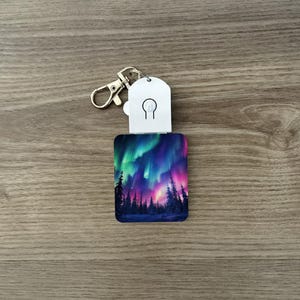 May include: A rectangular keyring featuring an aurora borealis design. The image shows a colourful display of green, purple, and pink lights above a forest silhouette. The keyring has a silver clasp and a white tag with a lightbulb icon.