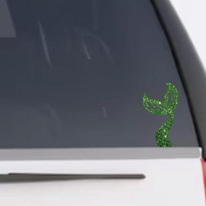 May include: A green glittery mermaid tail decal applied to the rear window of a vehicle. The decal features a textured, sparkling finish and a curved, bifurcated tail design that is positioned vertically on the glass surface.