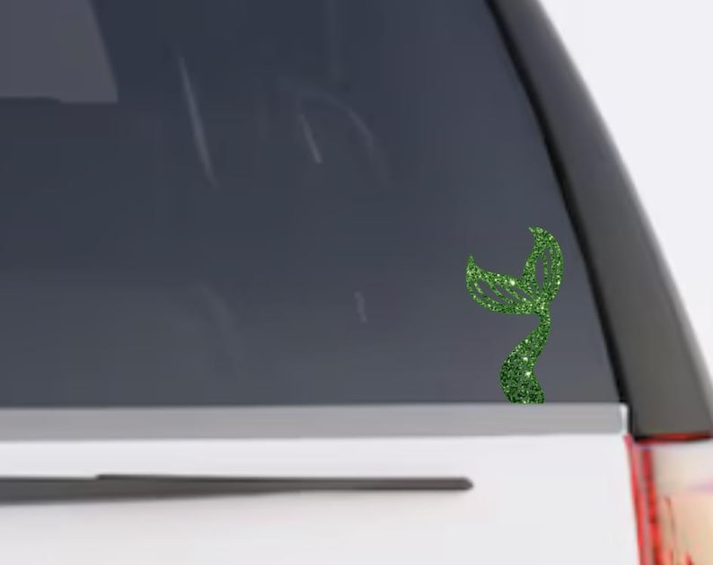 May include: A green glittery mermaid tail decal applied to the rear window of a vehicle. The decal features a textured, sparkling finish and a curved, bifurcated tail design that is positioned vertically on the glass surface.