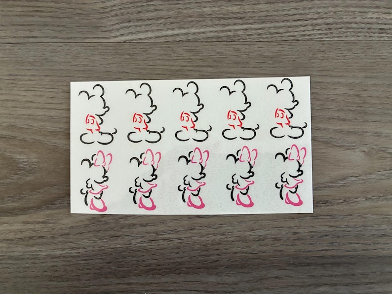 May include: A sheet of white stickers featuring cartoon character outlines. The top row has black and red images, while the bottom row has black and pink images. The stickers are arranged in a grid pattern on a white rectangular sheet.