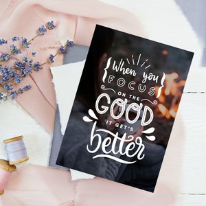 May include: A black and white inspirational quote card with the text "When you focus on the good it gets better" in a white handwritten font. The background image is a blurry photo of a person holding a mug in front of a campfire.