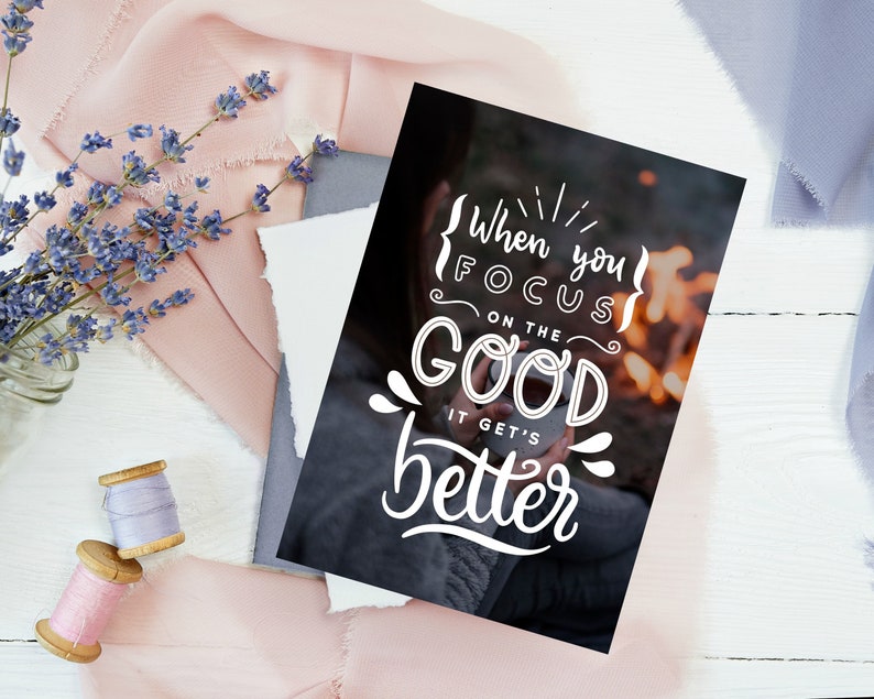 May include: A black and white inspirational quote card with the text "When you focus on the good it gets better" in a white handwritten font. The background image is a blurry photo of a person holding a mug in front of a campfire.