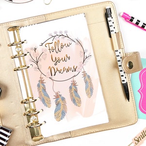 May include: A gold binder with a white page open to a page with the text "Follow Your Dreams" in gold lettering. The page has a pink and white watercolour background with a dreamcatcher design with feathers.