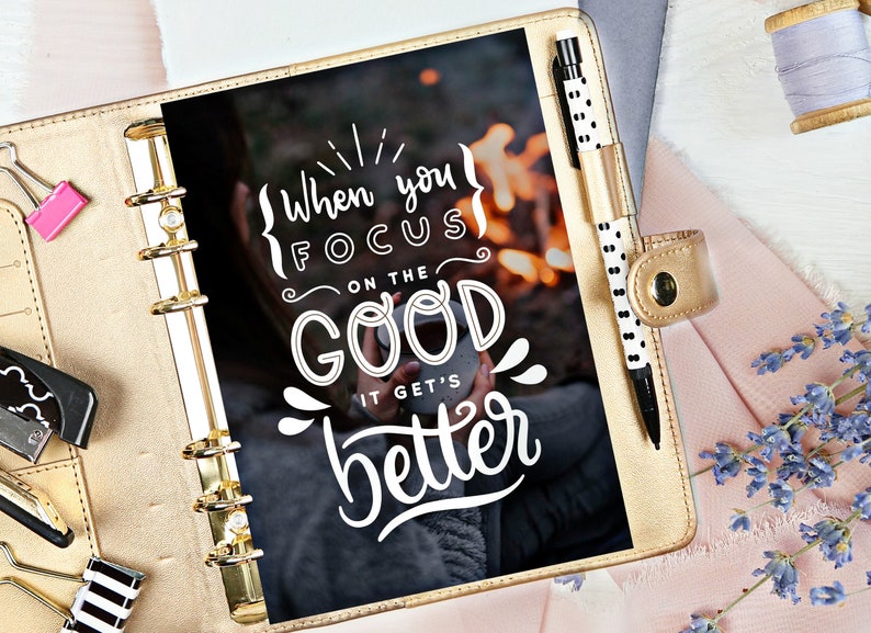May include: A gold-coloured planner with a black page featuring the text "When you focus on the good it gets better". The page has a blurry image of a person holding a mug in front of a campfire.