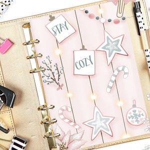 Puede incluir: Una agenda rosa y blanca con anillas doradas. La agenda tiene una variedad de pegatinas, incluyendo estrellas, bastones de caramelo y un adorno navideño. La agenda también tiene las palabras "Stay Cozy" escritas en ella.