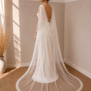 May include: A floor-length ivory bridal cape made of sheer tulle fabric. The cape features a delicate scalloped lace trim along the edges and attaches at the shoulders, draping down the back to create a long, flowing train over a simple gown.