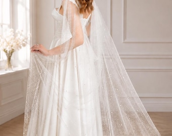 May include: A floor-length white gown featuring a sweetheart neckline and a long, sheer, sparkling cape attached at the shoulders. The cape is made of a translucent, glittery fabric adorned with small, scattered white pearls that trail to the floor.