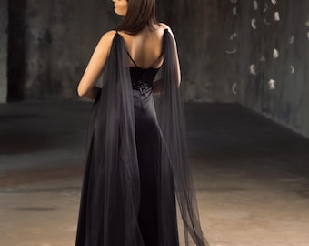 May include: A long, black formal gown with sheer, flowing cape sleeves. The dress features a fitted bodice and a floor-length skirt. The model is facing away from the camera, showcasing the back of the dress. The background is a dark, textured wall.