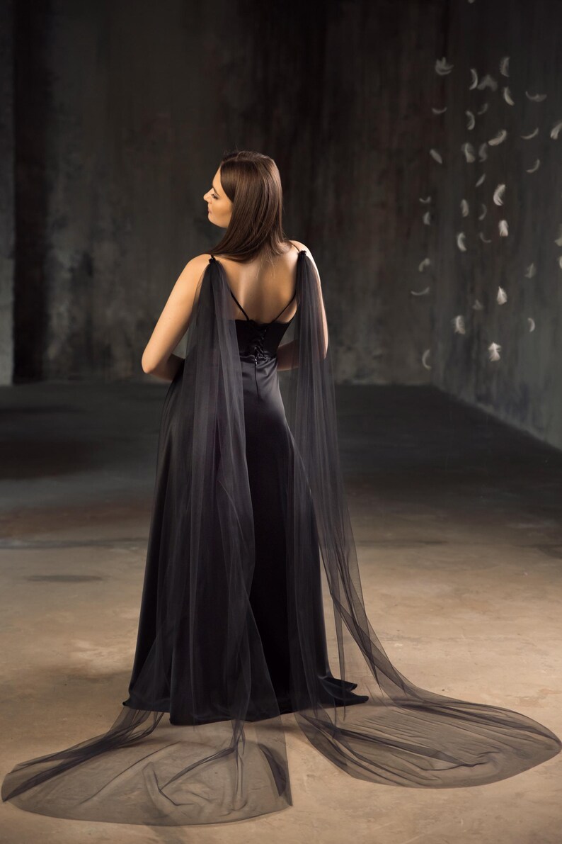 May include: A floor-length black gown featuring a fitted bodice with thin spaghetti straps and a corset-style back closure. The dress includes long, sheer black tulle panels attached at the shoulders that drape down to form a flowing train.