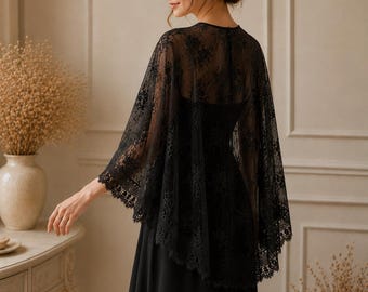 May include: A black lace capelet featuring an intricate floral pattern and scalloped edges, worn over a solid black sleeveless dress. The sheer, lightweight fabric drapes over the shoulders, providing a delicate overlay with a detailed hem.