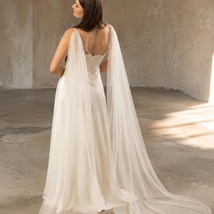 May include: A floor-length ivory satin dress featuring a lace-up back closure and long, sheer tulle cape sleeves that drape from the shoulders to the floor, creating a flowing silhouette with a train that extends behind the garment.