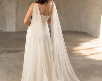 May include: A floor-length ivory satin dress featuring a lace-up back closure and long, sheer tulle cape sleeves that drape from the shoulders to the floor, creating a flowing silhouette with a train that extends behind the garment.