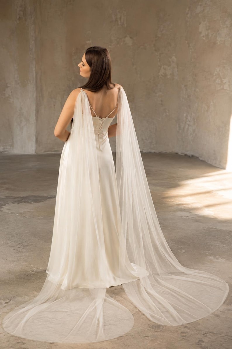 May include: A floor-length ivory satin dress featuring a lace-up back closure and long, sheer tulle cape sleeves that drape from the shoulders to the floor, creating a flowing silhouette with a train that extends behind the garment.