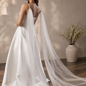 May include: A floor-length white satin gown featuring a square neckline and thin spaghetti straps. Attached to the straps are long, sheer, flowing tulle panels that drape down the back, extending onto the floor to create a dramatic train effect.