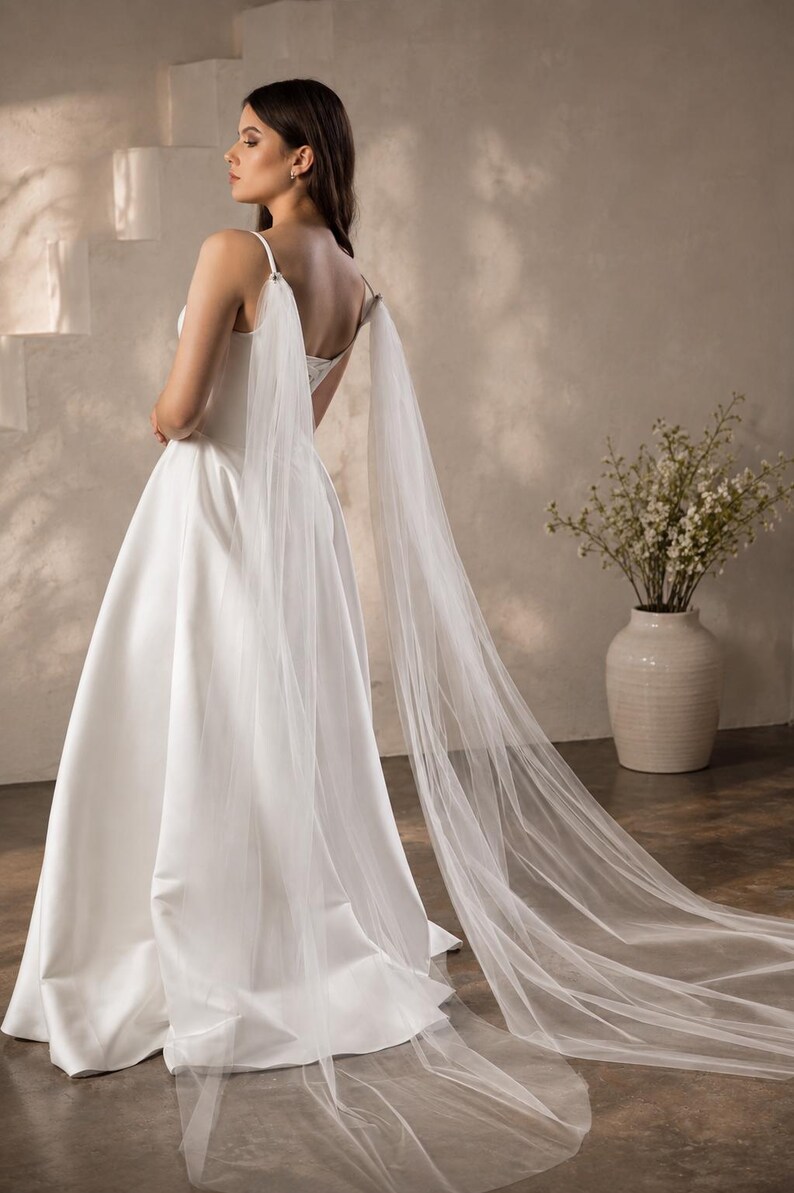 May include: A floor-length white satin gown featuring a square neckline and thin spaghetti straps. Attached to the straps are long, sheer, flowing tulle panels that drape down the back, extending onto the floor to create a dramatic train effect.