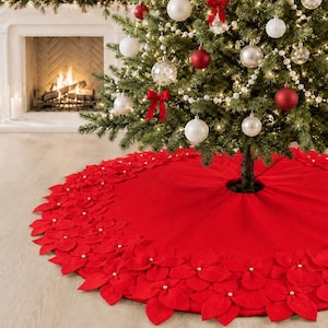 May include: A circular red Christmas tree skirt featuring a decorative border of layered felt poinsettia petals. Each fabric flower is accented with a small white bead at its center. The skirt has a central opening for the tree trunk.