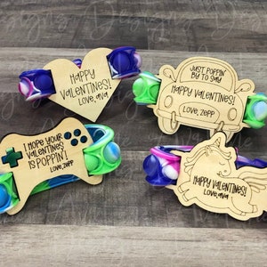 Puede incluir: Cuatro pulseras de madera para el Día de San Valentín con juguetes de burbujas pop-it adjuntos. Las pulseras presentan diferentes diseños, que incluyen un corazón, un coche, un mando de videojuegos y un unicornio. Cada pulsera tiene un mensaje personalizado, como "¡Feliz Día de San Valentín!" y "¡Espero que tu Día de San Valentín esté genial!".