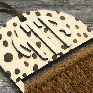May include: A wooden wall hanging with a brown and white cheetah print design. The hanging features a large monogram with the letters "MTP" and a long brown tassel.