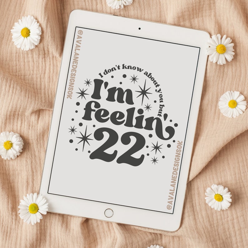 May include: A white tablet screen with a black graphic design that says "I don't know about you but I'm feelin' 22" with stars and dots around the text. The tablet is on a beige textured surface with white flowers.