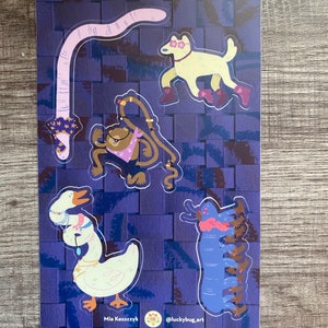 Op de afbeelding: Vijf kleurrijke stickers met een roze worm, een witte hond met een zonnebril, een bruine kikker met een zonnebril en een bandana, een witte gans met een zonnebril en een ketting, en een blauwe duizendpoot. De stickers zijn op een paarse achtergrond met een wit rasterpatroon. De tekst "Mia Keszczyk" en "@luckybug.art" staat onderaan de afbeelding.