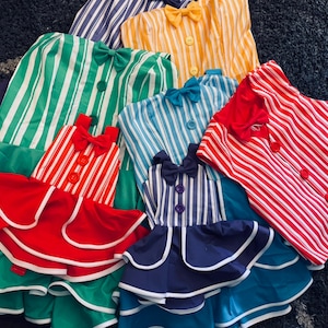 May include: Five striped dresses in various colors, including red, green, blue, and yellow. Each dress has a bow at the neckline and a button closure. The dresses are layered on top of each other, creating a colorful and playful display.